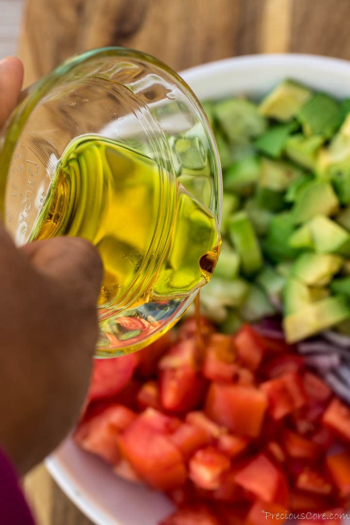 Hand holding a bowl of olive oil and pouring over avocado salad.
