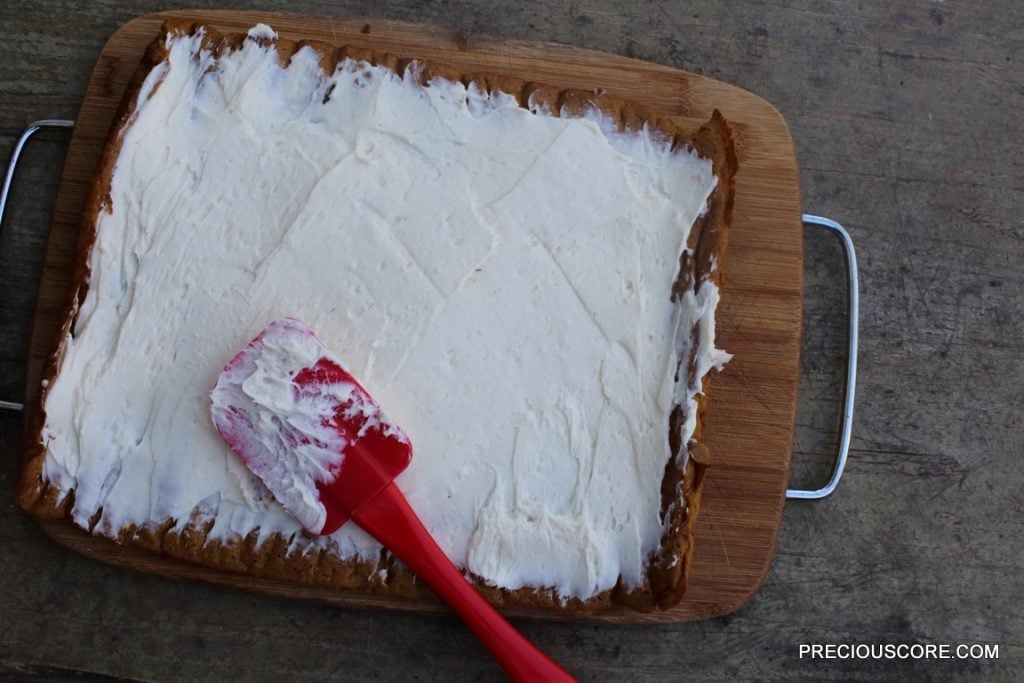 Red spatula spreading cream cheese filling onto pumpkin cake.