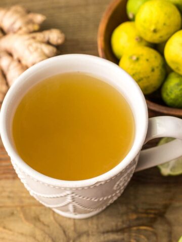 A mug of fresh ginger drink with some ginger and key limes on the side