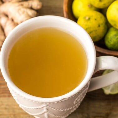 A mug of fresh ginger drink with some ginger and key limes on the side