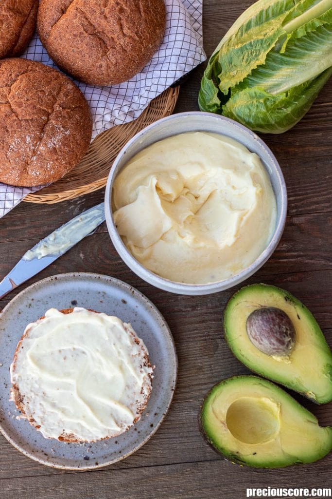 homemade mayoyonnaise in a bowl, bread buns in a basket, half a bun with mayo spread on top, surrounded by lettuce and avocado.