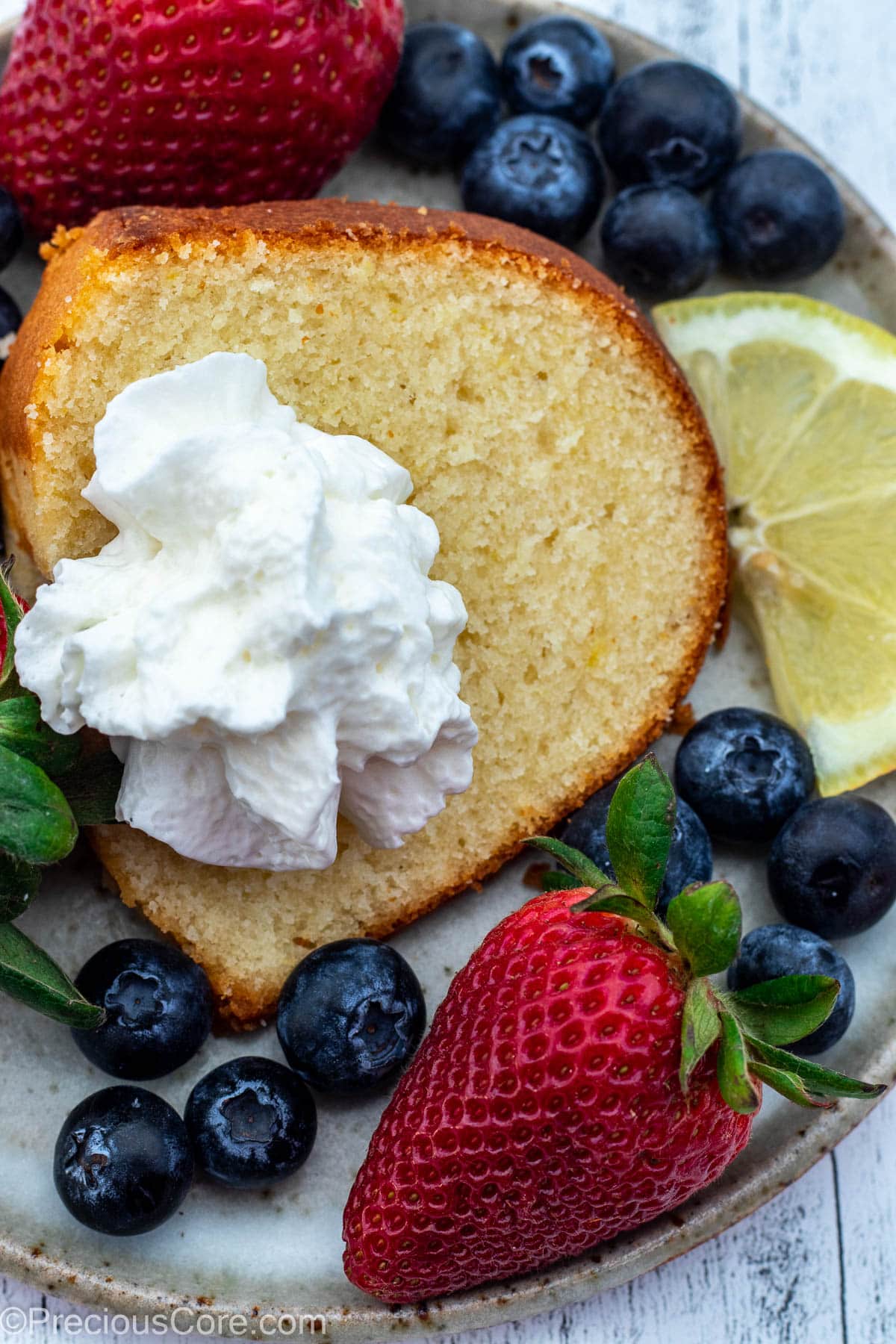 A slice of lemon pound cake on a plate, topped with whipped cream and surrounded by berries.