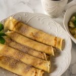 repes on a plate with a cup of coffee and a bowl of fruits nearby