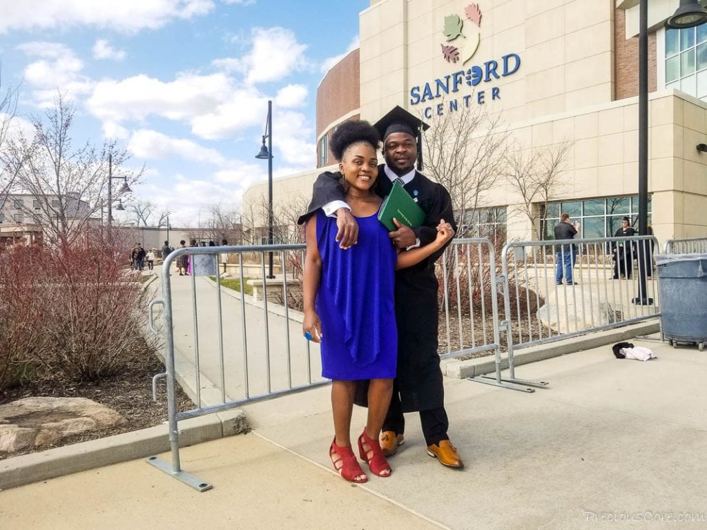 Man in graduation gear with a woman in a blue dress in front of the Sanford Center.