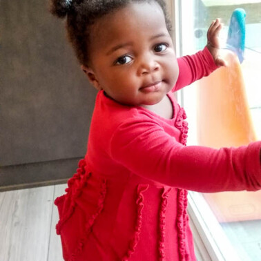 Baby in a pink dress standing at a window, looking at the camera.