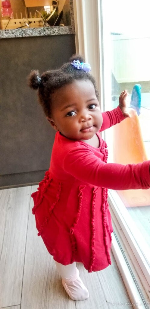 Baby in a pink dress standing at a window, looking at the camera.