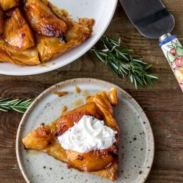 Slice of tarte tatin on a plate topped with cream, plate of large tarte tatin in the back