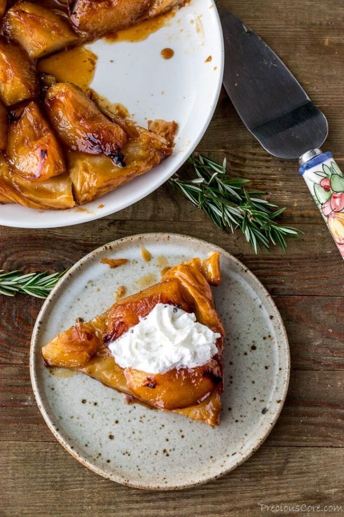 Slice of tarte tatin on a plate topped with cream, plate of large tarte tatin in the back