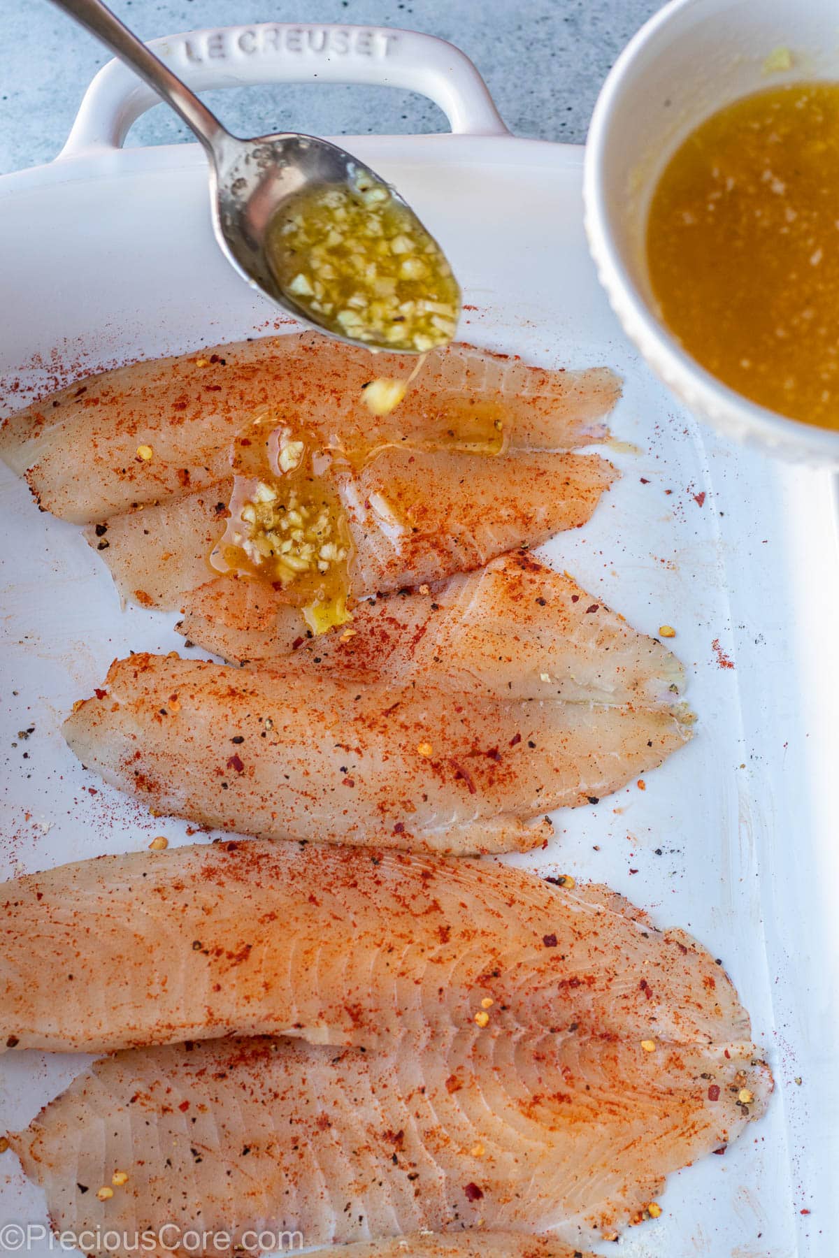 Spoon pouring garlic butter sauce over seasoned tilapia fillets.