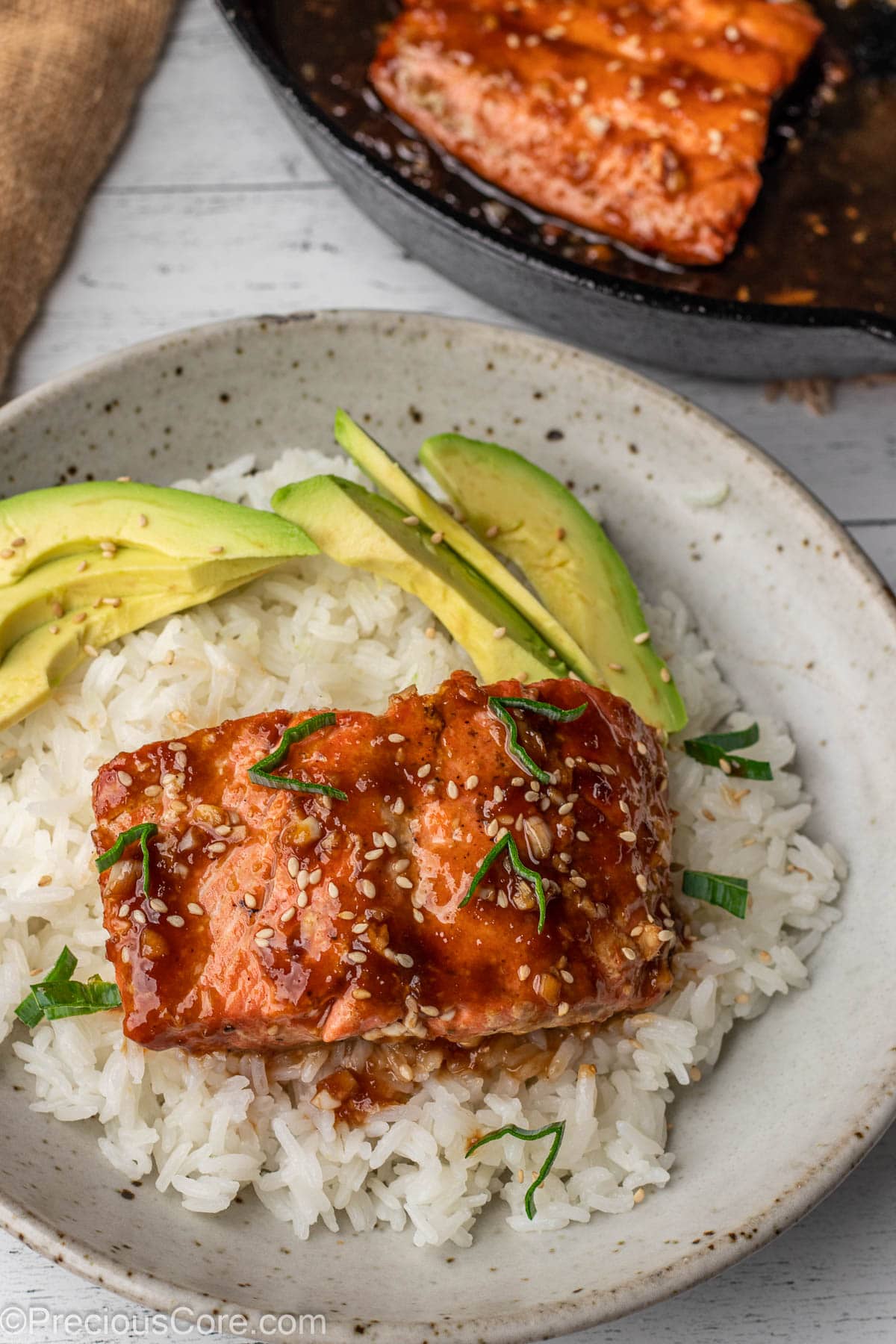 Piece of teriyaki salmon over white rice in a bowl, with avocado slices on the side.