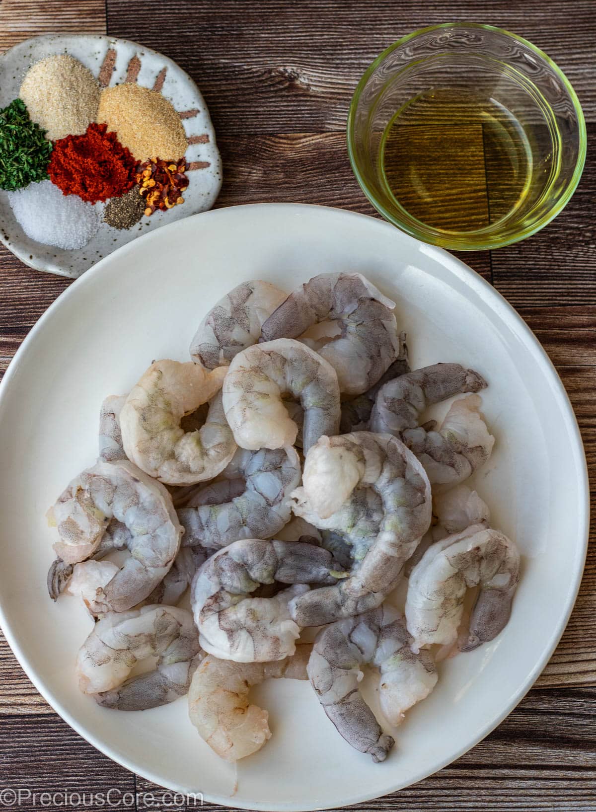 Raw shrimp on a plate, dried seasoning on a plate and olive oil in a small bowl.