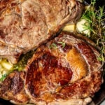 Two ribeye steaks in a cast iron skillet with rosemary, thyme, and garlic.