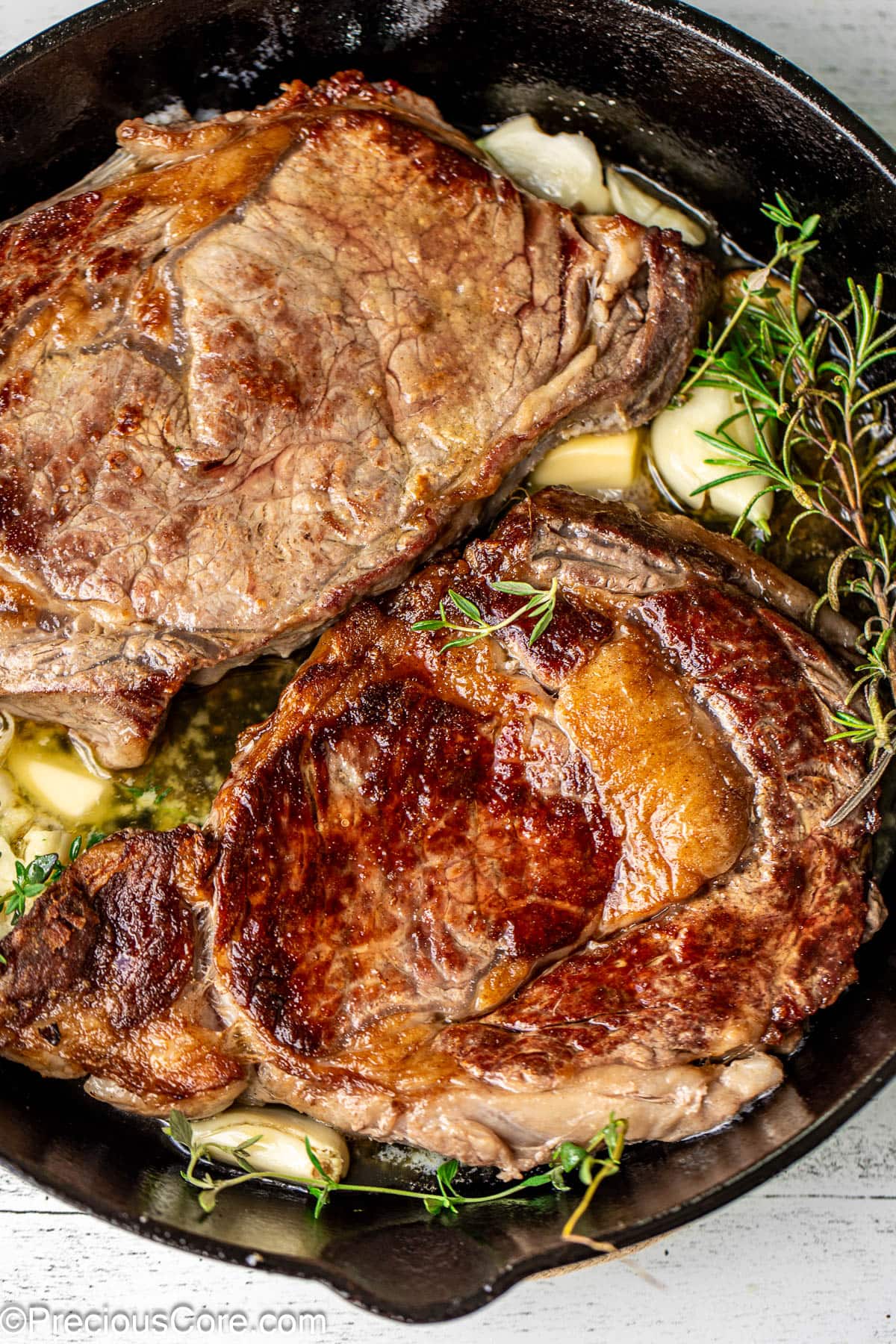 Two ribeye steaks in a cast iron skillet with rosemary, thyme, and garlic.